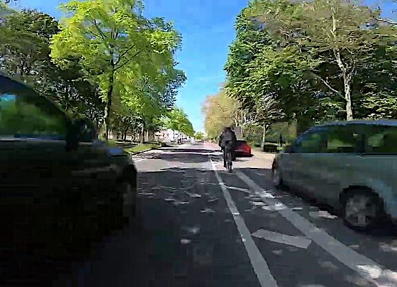 Ein Fahrradfahrer fährt neben einem zugeparkten Radstreifen, während sich ein weiteres Kfz von hinten nähert