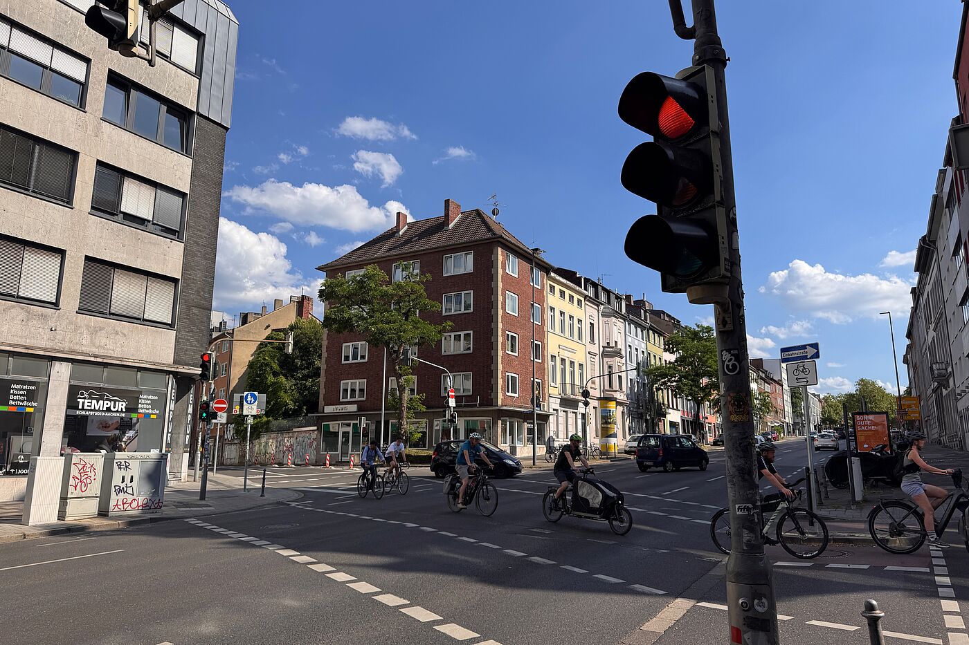 Mehrere Menschen auf Fahrrädern und einem Lastenrad queren eine Kreuzung an der Wilhelmstraße. Die Fahrbahn ist breit, Ampeln und Kfz-Verkehr prägen die Situation.