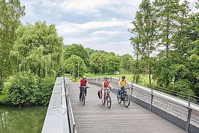 Drei Radfahrende befinden sich auf einer großen hölzernen Fahrrad- und Fußgängerbrücke. Im Hintergrund sind Bäume und eine  Graslandschaft zu sehen
