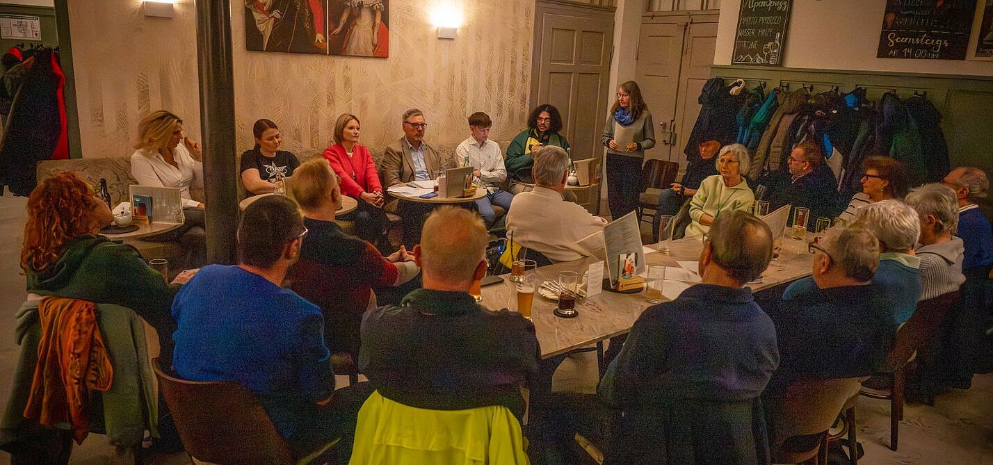 Landtagskandidat*innen sitzen nebeneinander auf einer Bank im Kaffeehaus, davor aufmerksam zuhörende Menschen aus dem Publikumtgliedern und Interessierten im Kaffeehaus Gutmann