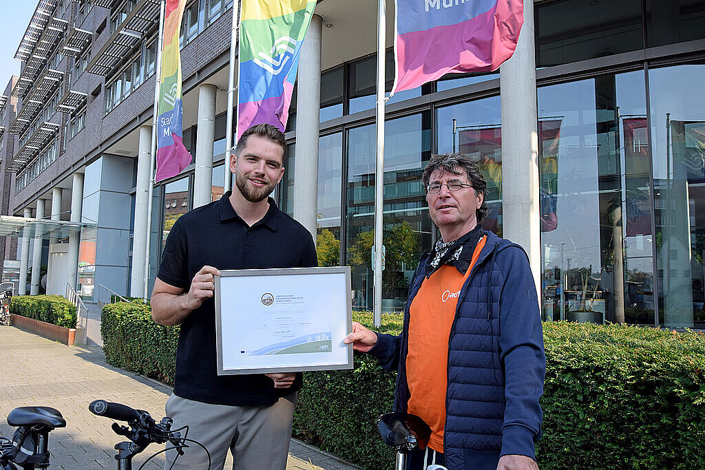 Zwei Männer stehen vor einem Bürogebäude. Der rechts stehende Mann reicht dem links stehenden Mann ein eingerahmtes Zertifikat. Im Hintergrund wehen Fahnen in Regenbogenfarben. Im Vordergrund stehen zwei Fahrräder.