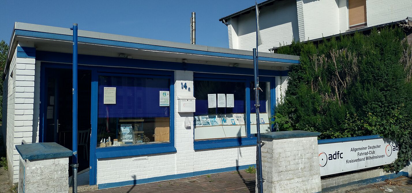 Infoladen Ein weißer, gemauerter Bungalow mit blauen Tür- und Fensterrahmen.