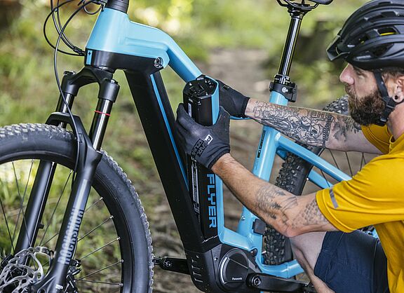 Ein Mann in gelbem Trikot und schwarzem Fahrradhelm entnimmt einen schwarzen Akku aus dem hellblauen Rahmen seines Pedelecs.