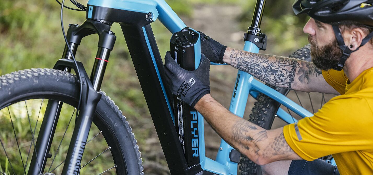 Ein Mann in gelbem Trikot und schwarzem Fahrradhelm entnimmt einen schwarzen Akku aus dem hellblauen Rahmen seines Pedelecs.